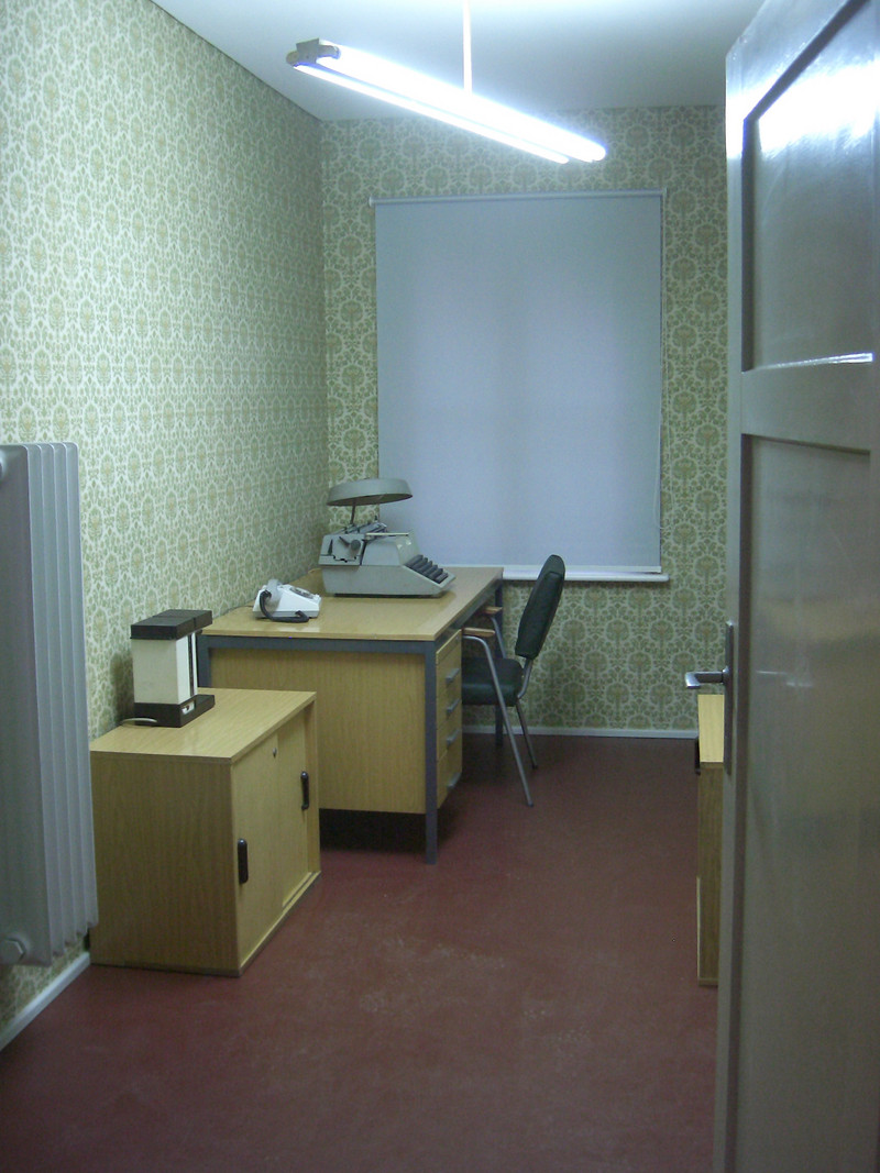 Das Foto rechts zeigt einen Verhörraum im Ausstellungsbereich MfS. Das Mobiliar – Schreibtisch mit Stuhl, Telefon und Schreibmaschine vom Typ „Optima“ sowie die Kaffeemaschine der Marke „Kaffeeboy S 600“ – stammt aus dem Fundus der Gedenkstä