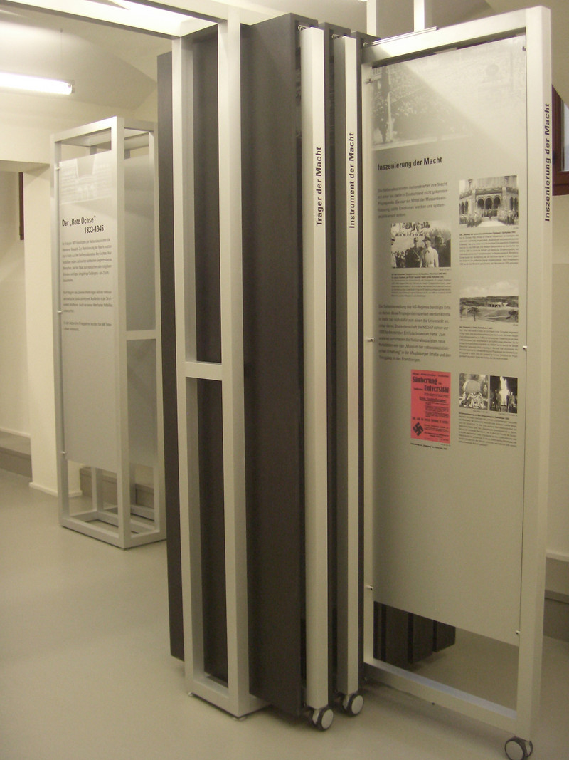 Das Foto links zeigt eines der Ausstellungselemente im Bereich Nationalsozialismus. Sie bestehen aus Tafeln mit Texten und Fotos zu einem speziellen Thema. Darüber hinaus können Flächen herausgezogen werden, die weitere Informationen enthalten.
